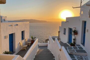 Traditional whitewashed houses with blue doors overlooking the Aegean Sea at sunset in Santorini, Greece – a peaceful Mediterranean alleyway bathed in golden hour light.