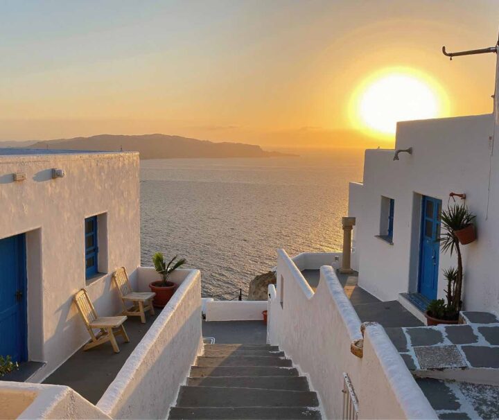 Traditional whitewashed houses with blue doors overlooking the Aegean Sea at sunset in Santorini, Greece – a peaceful Mediterranean alleyway bathed in golden hour light.