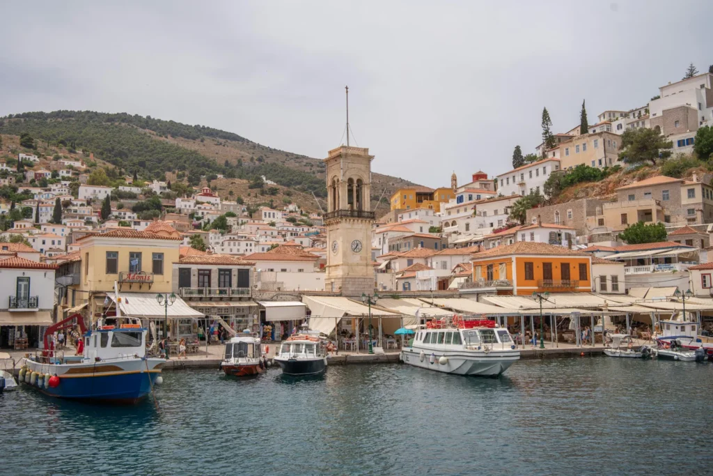 Harbor town with boats docked along a quay, colorful buildings, and a tall clock tower against a hillside.