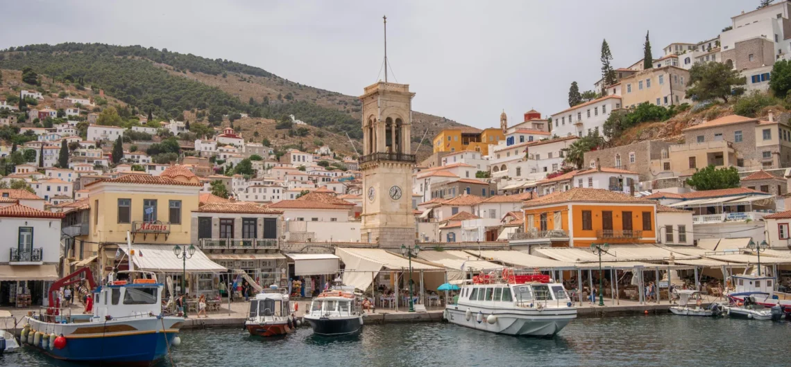 Harbor town with boats docked along a quay, colorful buildings, and a tall clock tower against a hillside.