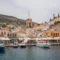 Harbor town with boats docked along a quay, colorful buildings, and a tall clock tower against a hillside.