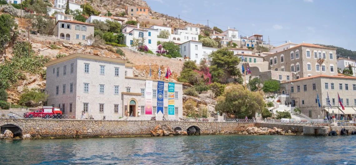 Coastal town with stone buildings along a rocky hillside and blue water in the foreground; banners hang from a waterfront building.