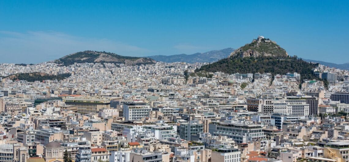 Panoramic cityscape of a dense Mediterranean town with two hills, clear blue sky, and white buildings sprawling across the scene.