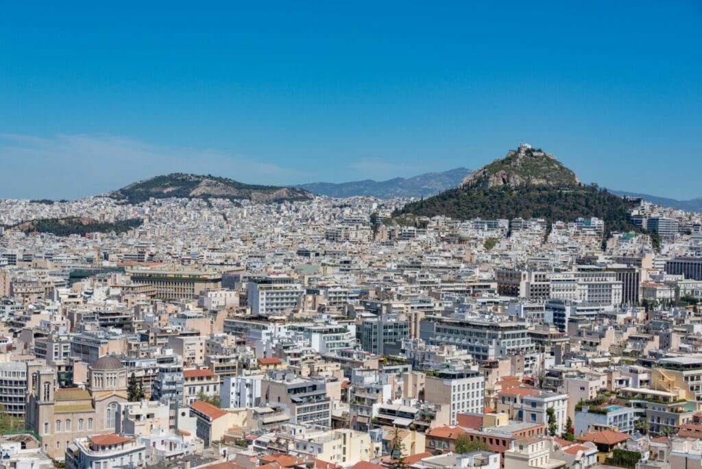 Panoramic view of a dense, low-rise city with two hills in the background, one with a rocky summit under a blue sky.