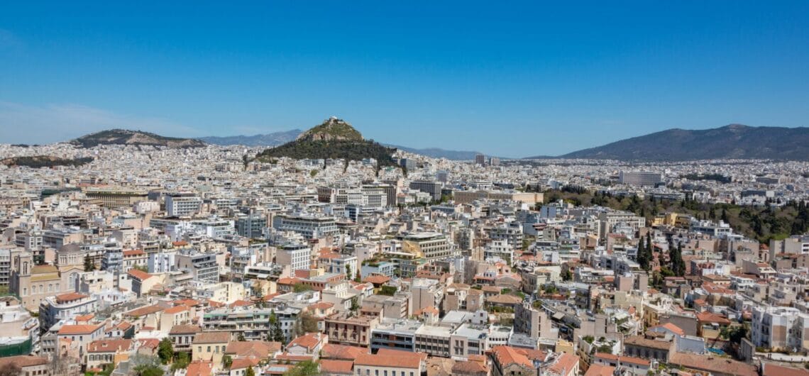 Wide cityscape of a densely built town with red-tiled roofs, low-rise and mid-rise buildings, and distant hills under a bright blue sky.
