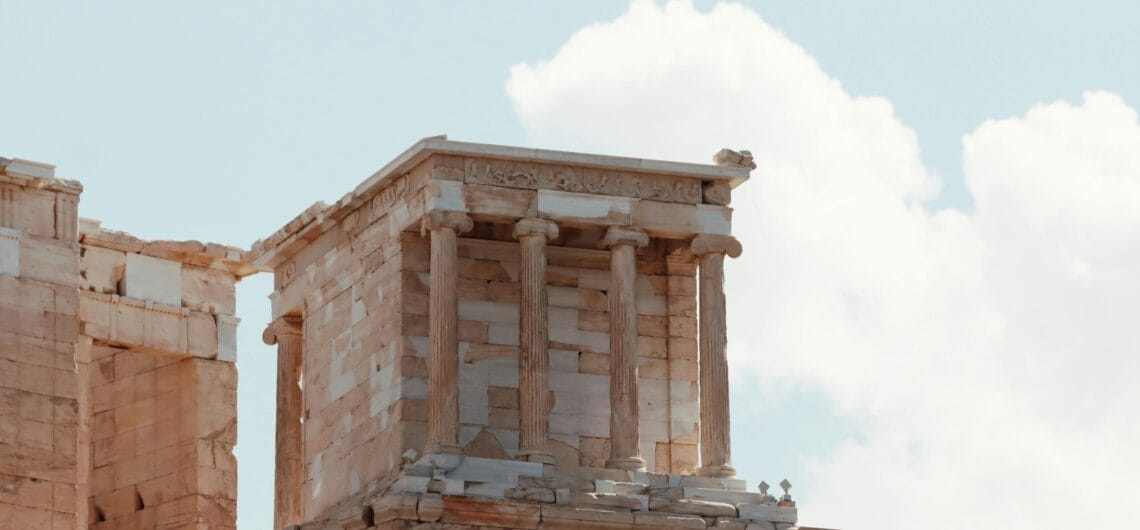 Ruined ancient stone temple facade with columns and carved frieze against a blue sky.