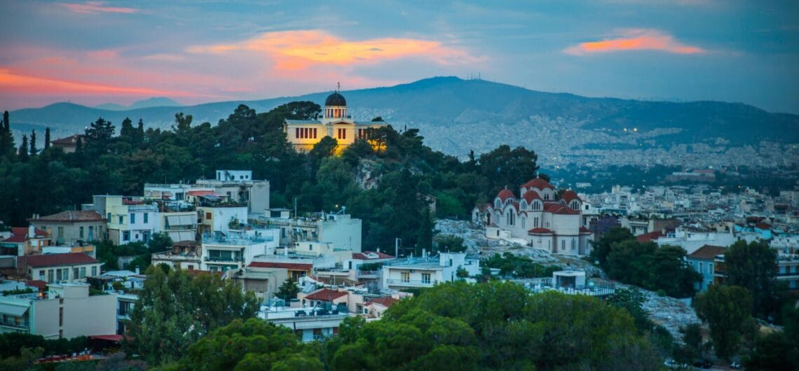 Cityscape of a hillside Greek town at dusk, with a domed hilltop church, white buildings, and lush trees under a blue-pink sunset sky.