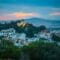 Cityscape of a hillside Greek town at dusk, with a domed hilltop church, white buildings, and lush trees under a blue-pink sunset sky.