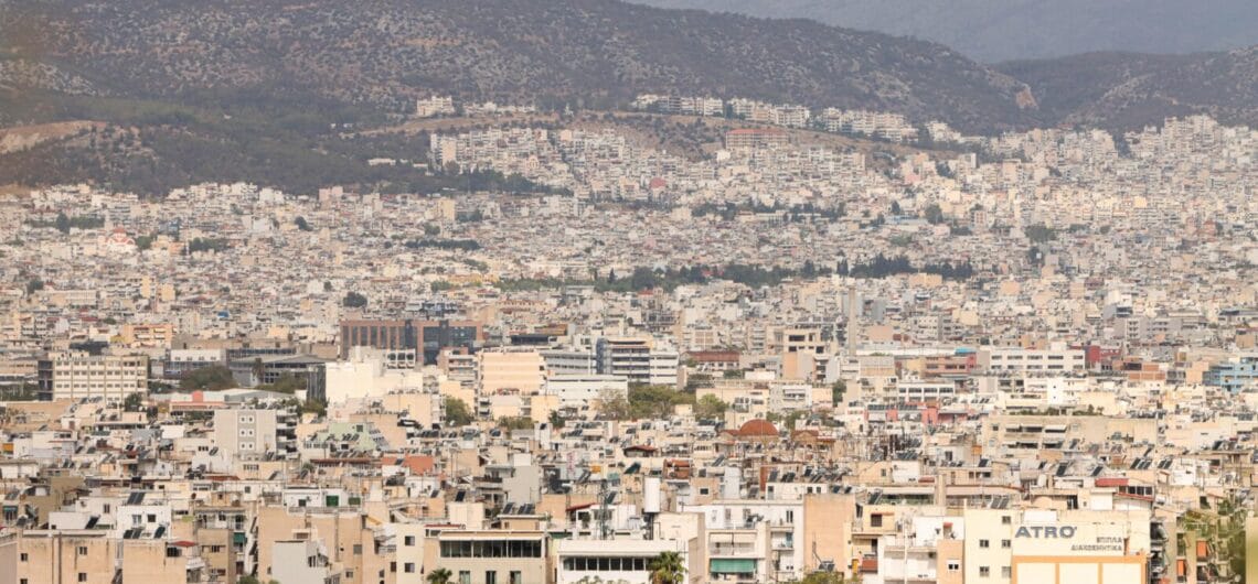 Dense cityscape sprawled across a valley with hills in the background and a sea of low-rise buildings in beige and white hues.