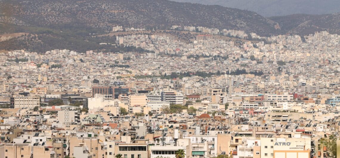 Wide view of a densely built city with beige apartment blocks stretching toward distant hills under a cloudy sky, hillside in the background.