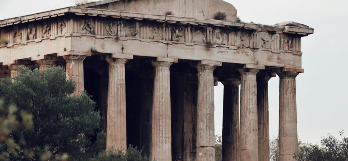 Ancient stone temple with tall columns partially hidden by bushes; visitors gather at its base.