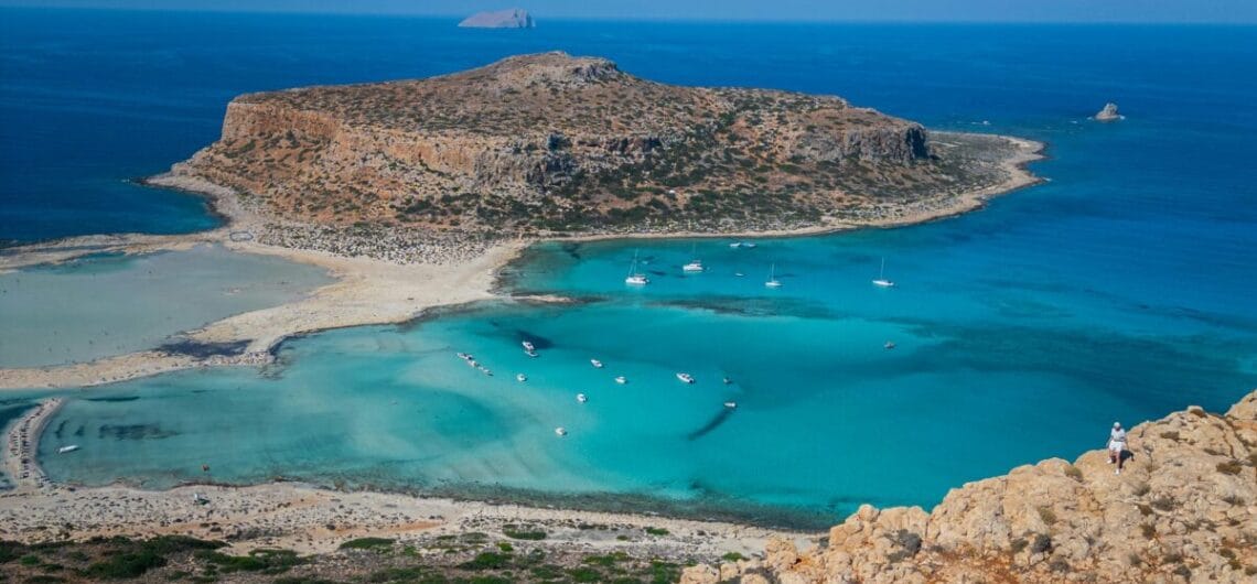 Rocky cliff and foreground overlook a turquoise bay with a sandy cove and several anchored boats on clear blue water.