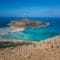 Rocky cliff overlooking turquoise sea with small boats in a sandy inlet and a distant island on the horizon in bright blue sky.