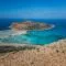 Rocky cliff overlooking turquoise sea with small boats in a sandy inlet and a distant island on the horizon in bright blue sky.