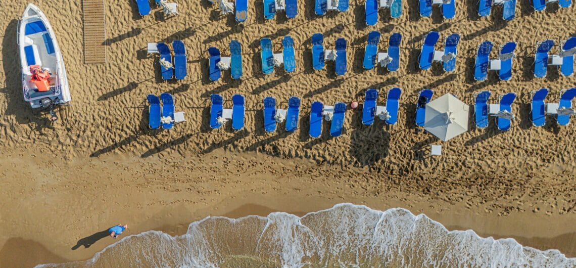 Aerial view of a crowded beach with dozens of blue lounge chairs arranged in neat rows, a boat on the left, and a wooden boardwalk.