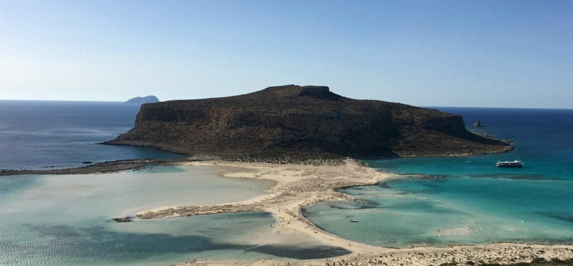 Rocky, flat-topped island with a sandy beach and turquoise shallow waters, boats anchored offshore nearby.