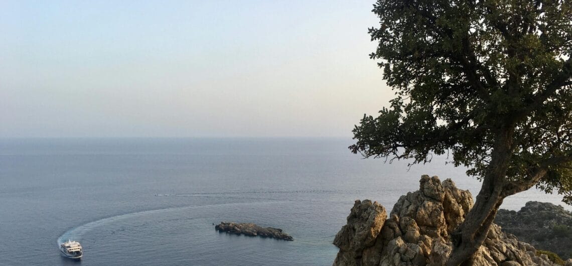 Coastline view with a rocky cliff and tree on the right overlooking calm sea; a small white boat near a rocky outcrop left of center.