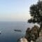 Coastline view with a rocky cliff and tree on the right overlooking calm sea; a small white boat near a rocky outcrop left of center.