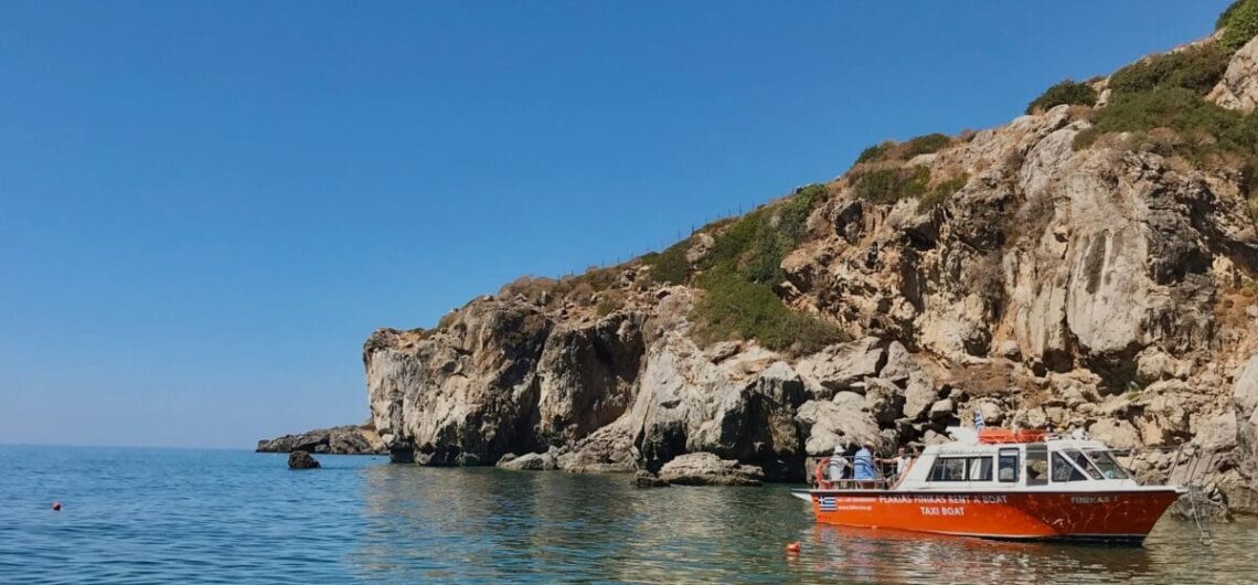 Red passenger boat near rocky shoreline, clear turquoise water and a pebbled seabed under a blue sky.