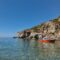 Red passenger boat near rocky shoreline, clear turquoise water and a pebbled seabed under a blue sky.