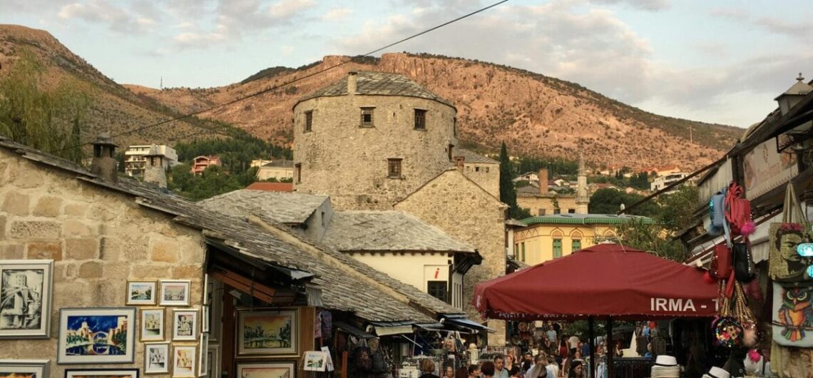 Stone-walled market street with art stalls and visitors, set against rocky hills and a blue sky.