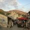 Stone-walled market street with art stalls and visitors, set against rocky hills and a blue sky.