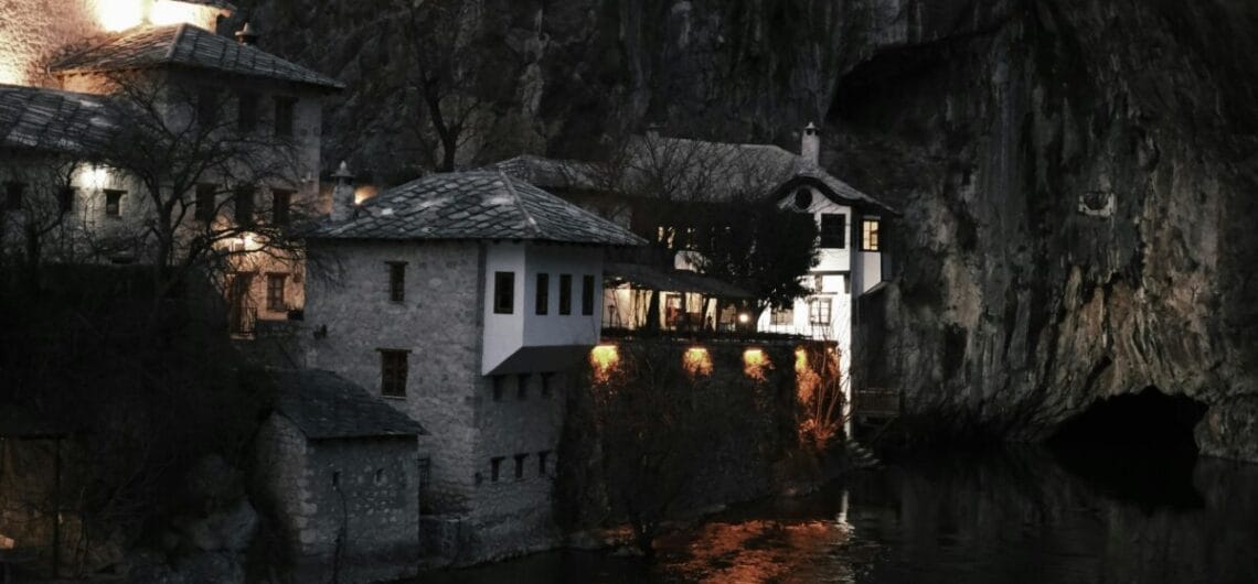 Stone houses line the riverbank at night, with a towering rocky cliff behind and warm lights reflecting on the water.