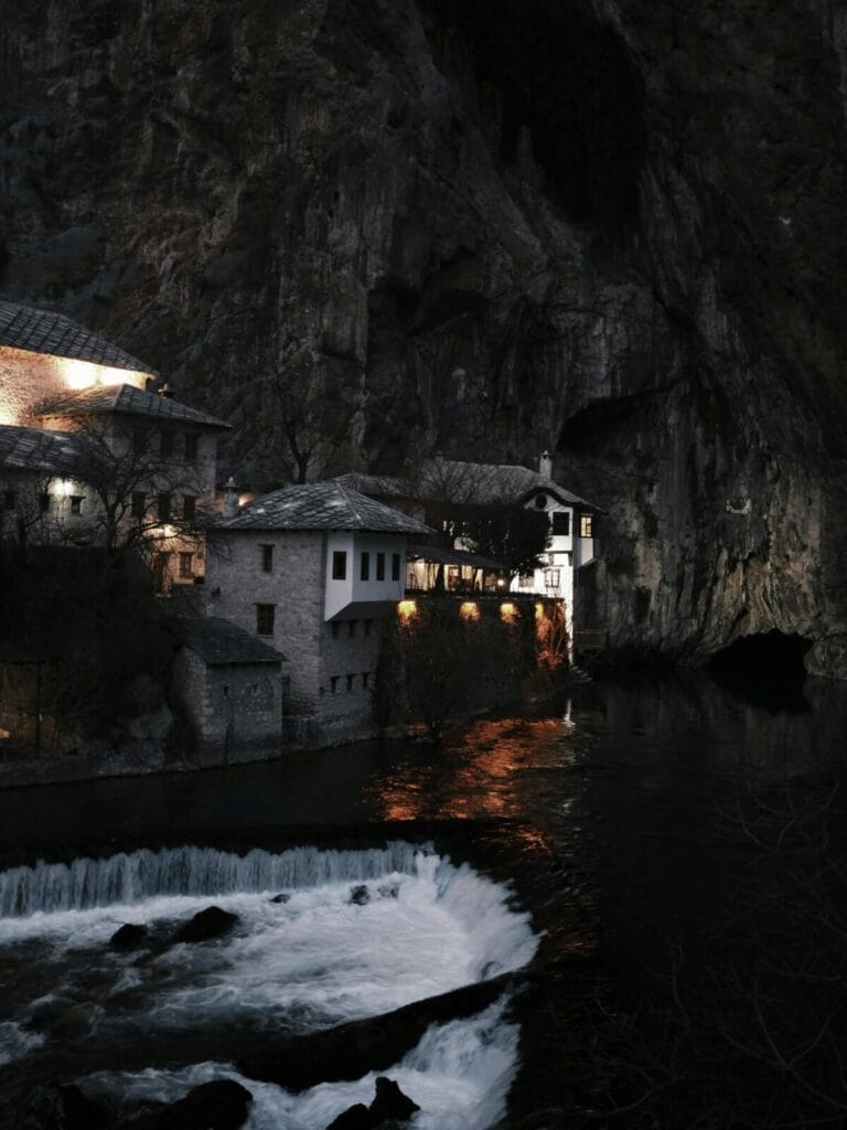Stone houses line the riverbank at night, with a towering rocky cliff behind and warm lights reflecting on the water.