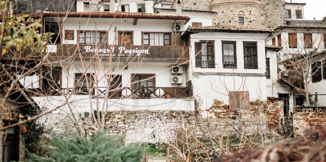 H hillside village inn with white plaster walls, stone accents and a wooden balcony, set among leafless trees and a garden.