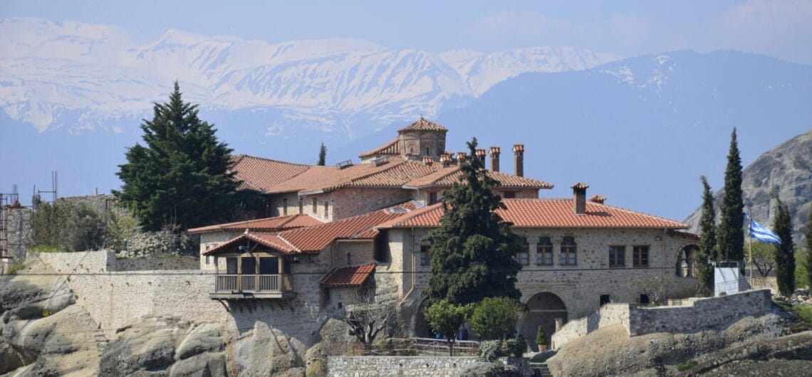 Stone villa with red-tiled roofs perched on rocky shore, with snow-capped mountains in the distance and a Greek flag to the right.