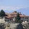 Stone villa with red-tiled roofs perched on rocky shore, with snow-capped mountains in the distance and a Greek flag to the right.