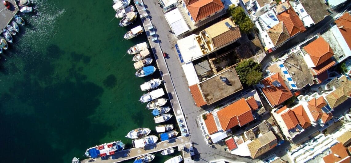 Aerial view of a harbor with boats docked along a quay and a town with orange-tiled roofs beside turquoise water.