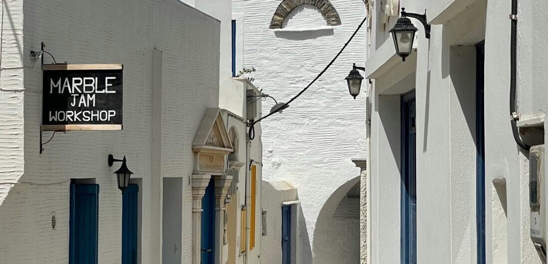 White-washed alley with blue doors and a sign reading Marble Jam Workshop, sunlit stone pathway.