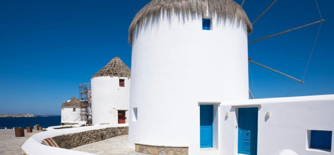 White cylindrical windmills with thatched roofs lining a stone promenade by the blue sea; vivid blue doors and windows in a sunny Mediterranean setting.