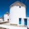 White cylindrical windmills with thatched roofs lining a stone promenade by the blue sea; vivid blue doors and windows in a sunny Mediterranean setting.