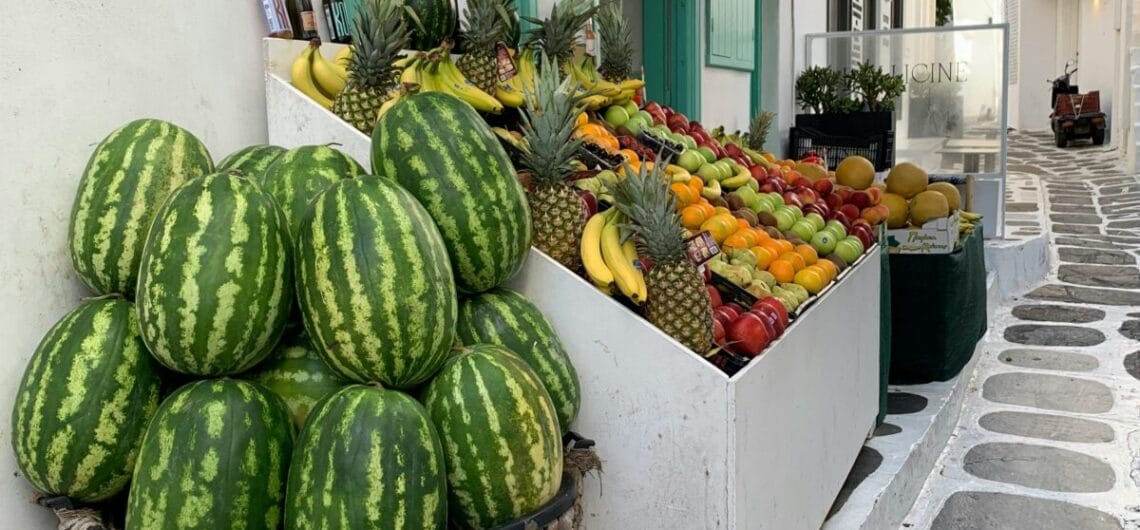 Baskets of watermelons and a colorful fruit stand outside a white-washed building with turquoise doors in a sunny Greek street.