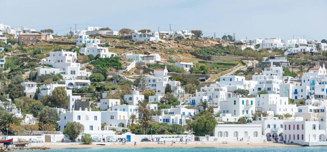 White-washed houses with blue doors cluster on a sunlit hillside overlooking a blue sea.