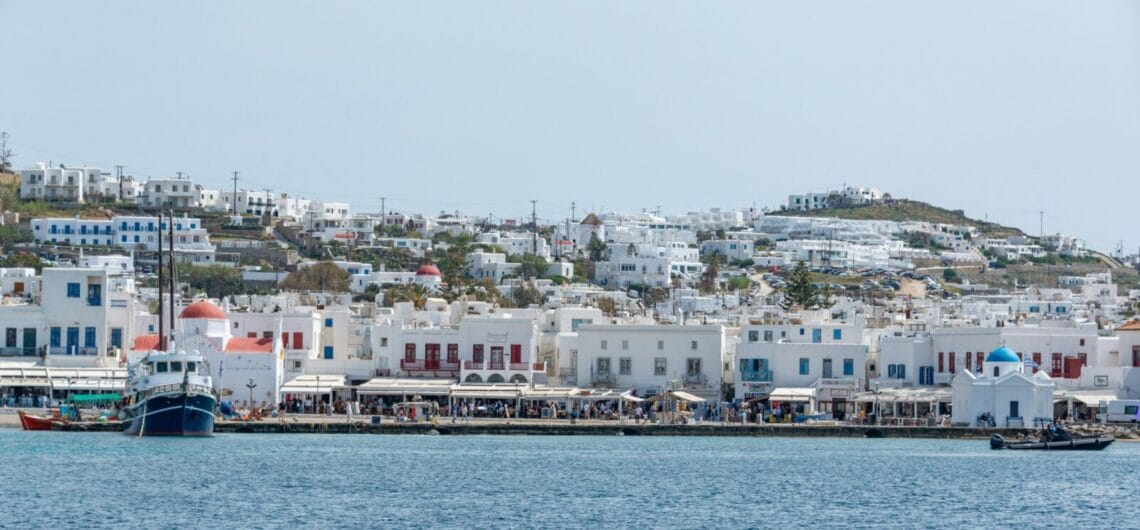 White-washed buildings with blue doors line the harbor in a Greek island town, boats docked along the quay.