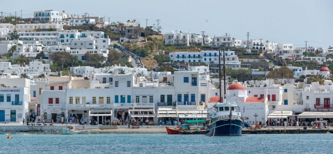 Coastal town with whitewashed buildings and blue doors along a harbor, calm blue sea in foreground and a large sailing ship docked by the waterfront.
