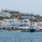 Coastal town with whitewashed buildings and blue doors along a harbor, calm blue sea in foreground and a large sailing ship docked by the waterfront.