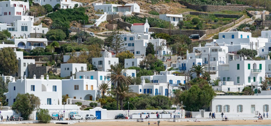 Whitewashed hillside town with blue doors and windows, terraced gardens, and a sandy beach by turquoise sea.