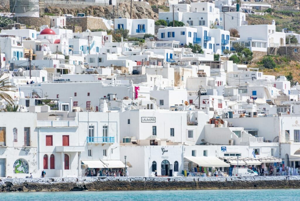 Whitewashed island buildings with blue doors and windows line a rocky waterfront, calm turquoise sea in the foreground (Santorini vibe).