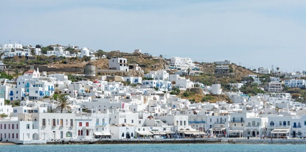 White-washed buildings cascade down a hillside to a calm blue sea under a pale sky, coastal town by the water.