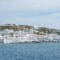 White-washed buildings cascade down a hillside to a calm blue sea under a pale sky, coastal town by the water.