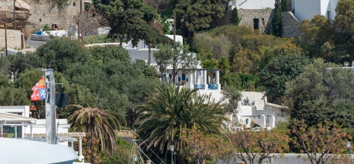 Whitewashed hillside village with blue doors and palm trees, a stone jetty, and a small harbor with a turquoise sea and a colorful boat.