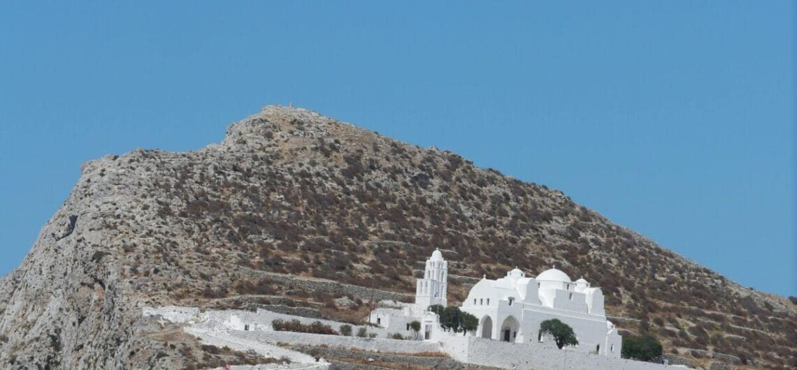 Whitewashed church with domes and a bell tower perched on a rocky hillside, with white-washed steps climbing the slope beneath a blue sky