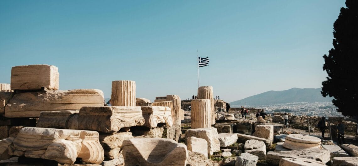 Ancient limestone ruins and stacked columns under a clear blue sky, with a Greek flag on a pole in the distance.