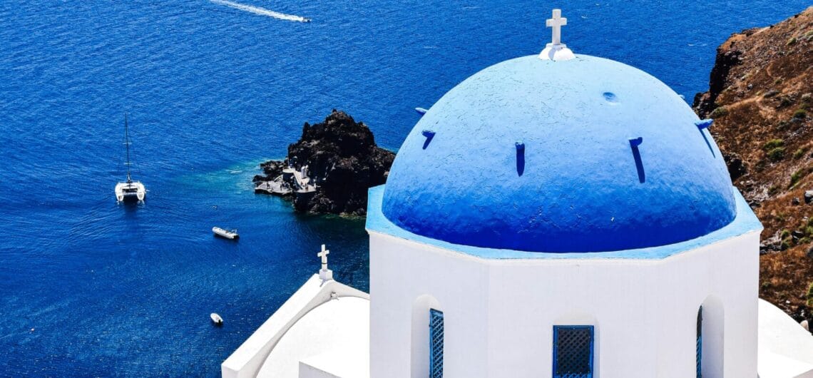 White Cycladic church with a blue dome and cross, overlooking the deep blue Aegean Sea and rocky cliffs above.