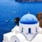 White Cycladic church with a blue dome and cross, overlooking the deep blue Aegean Sea and rocky cliffs above.