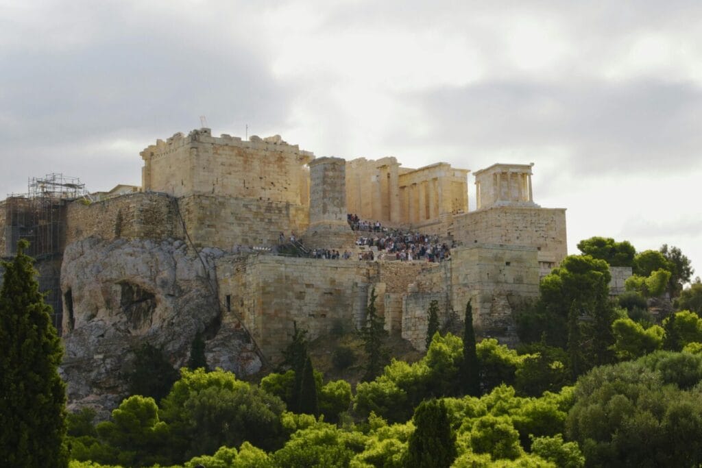 Ancient Acropolis ruins atop a rocky hill with visitors climbing the limestone steps, surrounded by trees beneath a cloudy sky.